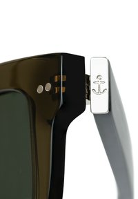 Gafas de sol con un marco cuadrado negro, acentos en marrón oscuro translúcido y un emblema de ancla plateado en la bisagra. Las lentes son de color verde oscuro.