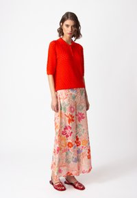 Red textured knit polo shirt with a collar, paired with a long floral skirt featuring pink, orange, and green patterns. Worn with red sandals.