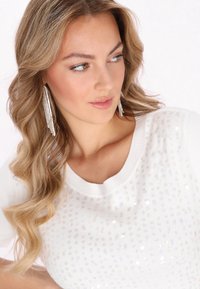 Weißer gestrickter Pullover mit rundem Halsausschnitt, verziert mit einem Muster aus silbernen Pailletten, die einen subtilen Glanz erzeugen. Getragen mit langen, metallischen Ohrringen.