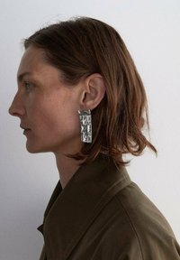 Orecchini geometrici in metallo argentato con forme rettangolari e quadrate, con una finitura lucida e texture, indossati da una persona con capelli di lunghezza media.