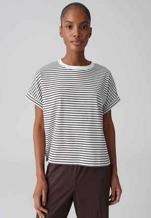 Vrouw draagt een wit shirt met korte mouwen en zwarte horizontale strepen, en donkere broek, staand tegen een effen lichte achtergrond.