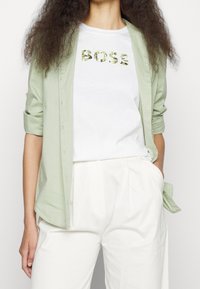 Camicia verde chiaro con bottoni sopra una t-shirt bianca con la scritta "BOSS" in foglie, abbinata a pantaloni color crema. Taglio semplice e rilassato.