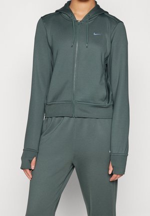 Osoba ubrana w ciemnozieloną bluzę z zamkiem i pasujące spodnie z otworami na kciuki oraz małymi logo Nike na klatce piersiowej i udzie.