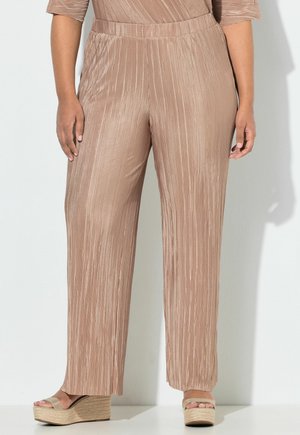 Vrouw die beige plissé wijde broeken met elastische tailleband en beige platformsandalen draagt, staande tegen een lichte achtergrond.