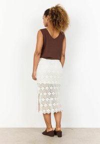 Haut sans manches marron, jupe midi blanche en dentelle avec un motif en zigzag, fente sur le côté, et chaussures compensées marron. Fond blanc uni.
