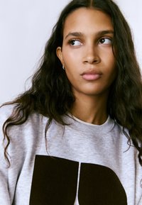 Sudadera gris con un llamativo estampado negro. El modelo tiene el cabello oscuro y ondulado, y lleva aros pequeños en las orejas. Toma cercana del rostro.