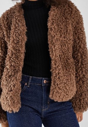 Veste en fausse fourrure bouclée marron portée sur un pull noir à côtes et un jean bleu.