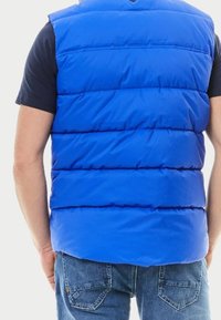 Blå vadderad väst med horisontell sömnad, rund halsringning och en slät, glänsande yta. Bärs över en mörk t-shirt, ihop med denimjeans.