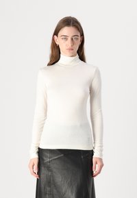 Pull col roulé en crème, fabriqué dans un tissu doux avec une silhouette ajustée. Manches longues et détail logo subtil au bas.