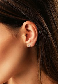 Nahaufnahme eines Ohres einer Frau mit glattem braunem Haar, die einen kleinen silbernen Stecker mit einem klaren Edelstein trägt.