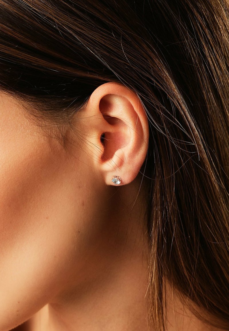 Nahaufnahme eines Ohres einer Frau mit glattem braunem Haar, die einen kleinen silbernen Stecker mit einem klaren Edelstein trägt.