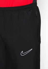 Zwarte sportbroeken met een elastische tailleband, voorzien van een witte geborduurde logo en een ritszak aan de zijkant, gemaakt van lichtgewicht stof.