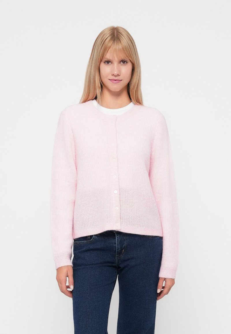 Rožinė megzta kardiganė su apvaliomis iškirpte, ilgomis rankovėmis ir mygtukų užsegimu. Minkšta tekstūra ir laisvas kirpimas, derinamas su tamsiai mėlynomis džinsomis.