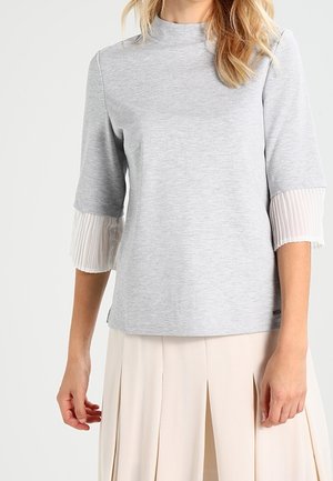 Gray long-sleeve top with a high neckline and pleated white cuffs, made of soft fabric, featuring a relaxed fit and minimalistic design.