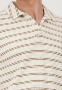 Camisa polo cremosa con finas rayas horizontales beige. Presenta un cuello clásico y una tela texturizada, ideal para el uso casual.