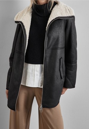 Zwarte shearling jas met crèmekleurige fleece voering, open gedragen over een zwarte coltrui, wit overhemd en beige broek.