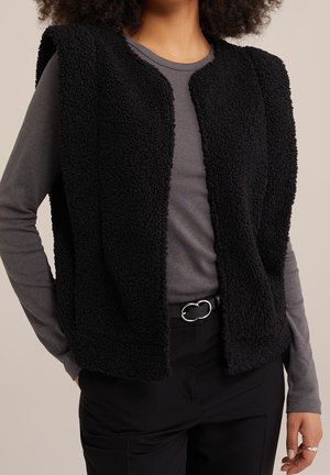 Kobieta w czarnej, teksturowanej, bezrękawnikowej kamizelce na szarym, długim rękawie, z czarnymi spodniami i podwójną sprzączką paska.