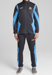 Chándal con una base negra y paneles laterales azules. Logos dorados en la chaqueta y los pantalones. El material parece liso y atlético.