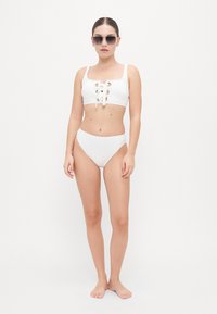 White bikini set featuring a cropped top with lace-up detailing and gold accents, paired with high-waisted bottoms. Smooth texture.