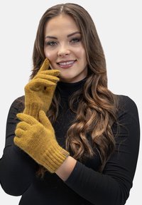 Sennepsgul strikkede handsker med ribbede manchetter, lavet af blødt materiale, har et mærke på håndleddet. Modellen poser med den ene hånd tæt på sit ansigt.