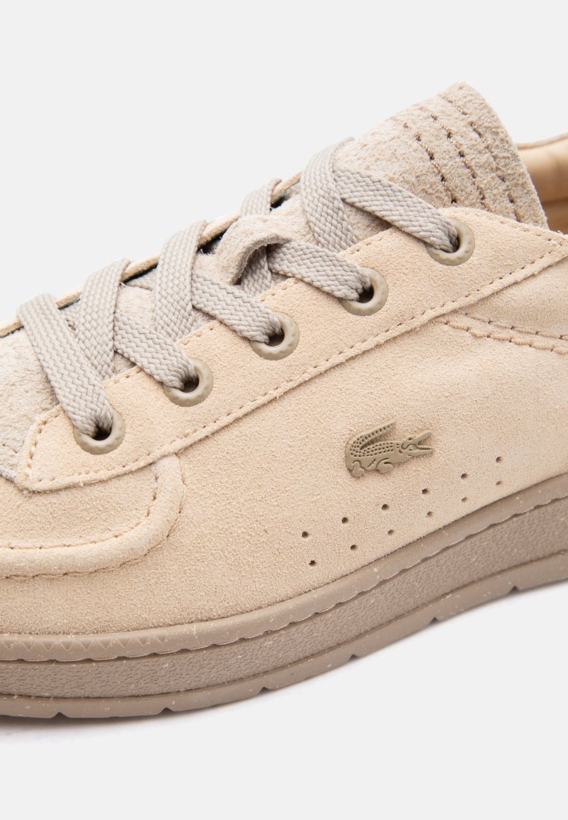 Close-up of beige suede sneaker with light grey laces, perforations near sole, and small crocodile logo on the side.
