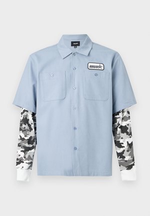Jeune homme aux cheveux bouclés portant une chemise bleu clair à manches courtes boutonnée par-dessus une chemise à manches longues imprimée camouflage noire, blanche et grise.