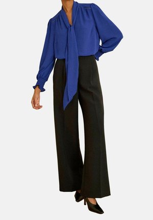 Femme portant un chemisier bleu à manches longues avec un nœud sur le devant, un pantalon noir à jambes larges et des escarpins noirs pointus à talons hauts.