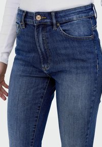 Mėlynos džinsinės kelnės su aukšta liemene, tiesiu kirpimu, priekinėmis kišenėmis ir klasikine mygtukų uždarymo sistema, su subtiliu išblukimu.