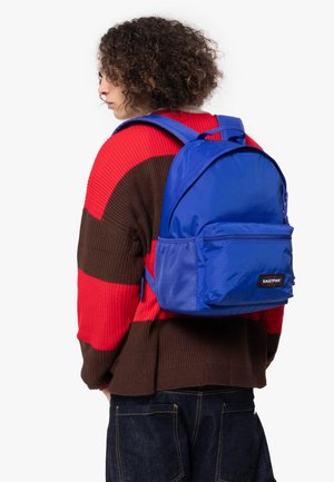 Blue nylon backpack with a front zip pocket and adjustable straps, worn over a red and brown striped sweater. Smooth, shiny texture.
