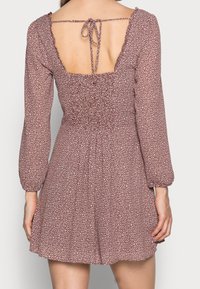 Mujer vistiendo un vestido mini de manga larga marrón con lunares blancos, que presenta una espalda cuadrada descubierta y un lazo en el cuello.