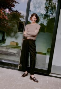Long-sleeve beige top paired with black pleated trousers and dark loafers. Smooth, textured materials with a braided tan belt accent.
