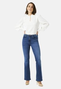 Vrouw staat met handen op de heupen, gekleed in een witte blouse met lange mouwen, blauwe high-waisted jeans en beige hoge hakken, met een glimlach.