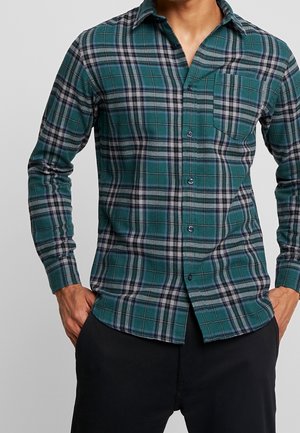 Homme portant une chemise à carreaux verte, noire et grise avec des boutons, les mains dans les poches d’un pantalon noir, sur un fond uni.