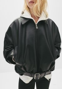 Svart läderjacka i oversized-modell med dragkedja fram, hög krage och bälte i midjan. Innehåller en grå hoodie under, vilket visar upp kontrasterande texturer.