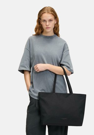 Sac fourre-tout noir avec des poignées robustes, présentant une texture lisse et un design minimaliste. Présenté sur un fond clair.