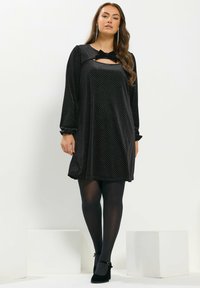 Schwarzes Kleid mit Polka-Dots, langen Ärmeln, einer Schleifen-Detail am Ausschnitt und entspannter Passform. Kombiniert mit schwarzen Strumpfhosen und geschlossenen High Heels.
