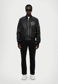 Giubbotto bomber in pelle nera con polsini a costine, chiusura con zip e logo circolare sul petto. Abbinato a pantaloni neri e sneakers bianche.