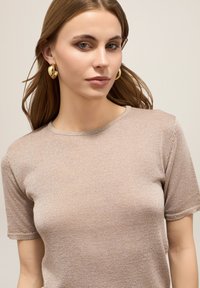 Beige korte mouwen top met een gebreide textuur en ronde hals, met een subtiele glans en omgeslagen zomen. Gouden oorringen maken de look compleet.