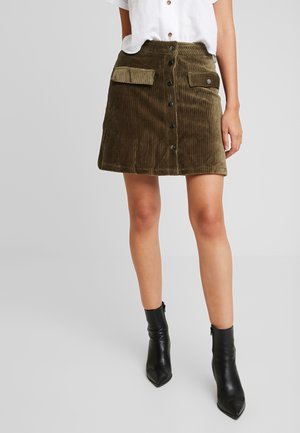 Olijfgroene corduroy rok met twee voorzakken, voorzien van een knoopsluiting aan de voorkant en getextureerde verticale strepen. Gecombineerd met zwarte enkellaarsjes.