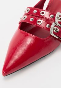 Scarpa rossa in pelle verniciata con punta affusolata, design a due cinturini con borchie e fibbie argentate, texture liscia e suola nera.
