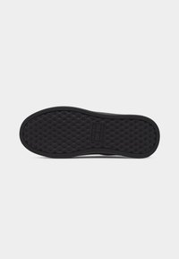Semelle de chaussure noire avec un motif antidérapant texturé et un détail de logo rectangulaire au centre sur un fond blanc.