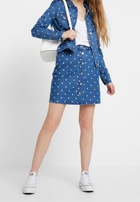 Woman wearing a polka dot denim jacket and skirt set, white V-neck shirt, white sneakers, and carrying a white shoulder bag.