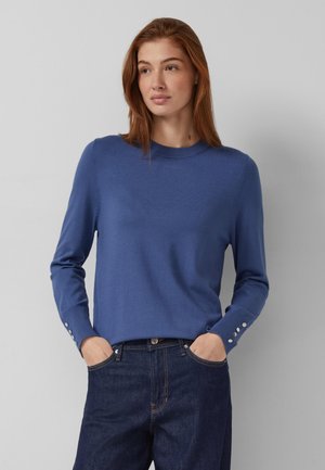 Sieviete ar sarkaniem matiem, tērpusies zilā džemperī ar pogām uz aprocēm un tumši zilās džinsās, rokām kabatās, skatās šķībi.