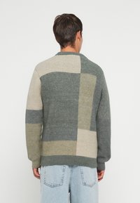 Maglione lavorato a maglia in diverse sfumature di verde e beige, caratterizzato da un design a patchwork con distinti motivi rettangolari e polsini a costina.