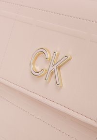 Rosa konstläderväska med sydda linjer, en framträdande guld- och elfenbensfärgad "CK"-logotyp samt en slät texturyta.