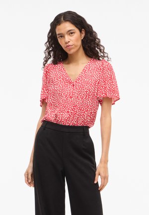 Γυναίκα με σγουρά μαλλιά φοράει κόκκινο μπλουζάκι με κοντά μανίκια και λευκά πουά, καθώς και μαύρο παντελόνι ψηλής μέσης, στέκεται μπροστά σε λευκό φόντο.