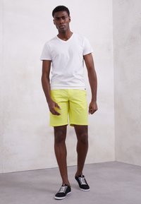 T-shirt bianco a V, pantaloni corti giallo brillante, scarpe da ginnastica nere. Tessuto liscio, design aderente e colori a contrasto. Il modello è in piedi contro uno sfondo neutro.