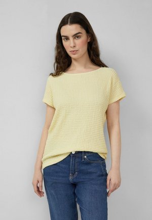 Frau mit langem braunem Haar, die ein strukturiertes, hellgelbes Kurzarmoberteil und blaue Jeans trägt, steht vor einem schlichten grauen Hintergrund.