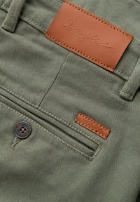 Tissu en coton vert olive avec une étiquette en cuir, comportant des coutures brunes et un détail de bouton noir. Comprend une poche arrière avec un accent en cuir.