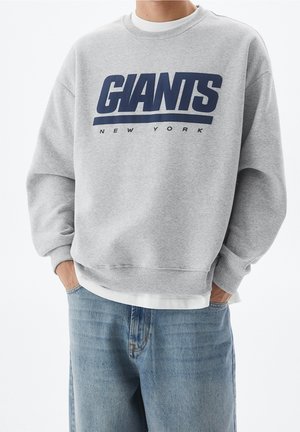 Osoba ubrana w jasnoszarą bluzę z granatowym napisem "GIANTS NEW YORK", w połączeniu z jasnoniebieskimi dżinsami, ręce w kieszeniach.